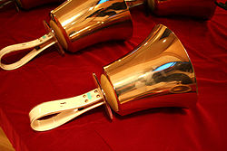 Zdjęcie przedstawia dwa dzwonki ręczne w kolorze złotym z uchwytami w kolorze białym. Dzwonki położone są obok siebie na czerwonym materiale.