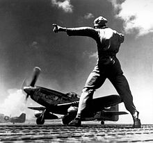 Black-and-white photograph of a man wearing military uniform facing a World War II-era single-engined fighter while gesturing to his left