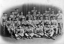 A formative photograph of white soldiers in African colonial-style uniforms, arranged in four rows; two standing, one sitting on chairs and one sitting on the floor