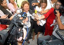 A distraught woman talks into a microphone with her eyes closed, surrounded by reporters. On the front of the woman's white T-shirt is a printed color portrait of a smiling man. Next to her stands a younger woman with her head down.