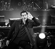 Photographie en noir et blanc de Jim Carrey en plein spectacle, il porte la main à son oreille comme pour mieux entendre