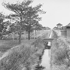 Black and white shot of a water-filled ditch, barbed-wire fences, and guard towers
