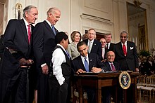 Photograph of Obama using a pen to sign a document, a crowd of people in suits behind him.