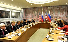 A conference room full of diplomats with US and Russian flags in the back against a wall.