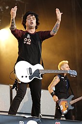 Two members of rock band Green Day shown onstage at a concert. From left to right, singer/guitarist Billie Joe Armstrong and bass guitarist Mike Dirnt. Behind them are a row of large guitar speaker cabinets. Billie Joe gestures with both hands to the audience.