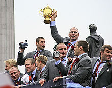 White men in grey suits, pale blue shirts and red ties celebrate upon the top floor of an open-top bus. On man holds a golden trophy in the air with one hand.