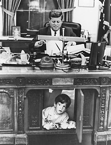 black and white image of John F. Kennedy seated at the Resolute desk with the center panel open and his young son playing in this opening.
