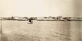 Photograph looking across water with a small sail boat in the foreground and buildings along the shore in the background