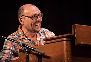 Bugge Wesseltoft wearing a light checkered shirt, sitting behind a piano, grinning broadly and looking left of camera