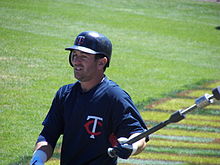 A man wearing a navy blue baseball jersey and batting helmet holds out a baseball bat in his left hand