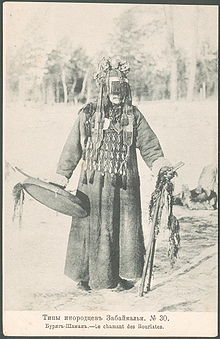 Faded black and white photo of a Mongol Buryat shaman, a masked man wearing a thick robe, an apron covering his chest, and a round hat with covering his face down to his nose. He is holding a drum in his right hand, and two decorated wooden sticks in his left hand. The caption in Russian and French means "Buryat shaman".