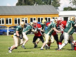 Players from two American football teams during a match. One team is wearing green shirts and gold pants, the other team is wearing red shirts with black pants
