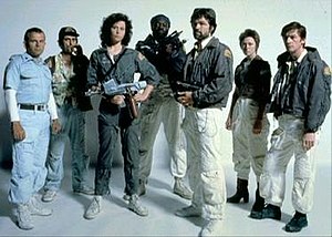 The seven principal cast members of Alien stand in front of a white backdrop, in costume and holding prop weapons from the film.