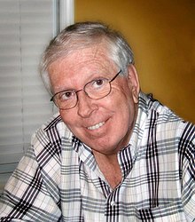 A middle-aged man with grey hair, wearing glasses and white shirt with black stripes