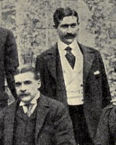 Sullivan, seated, with Herbert standing behind his left shoulder; both are very well-dressed and mustachioed